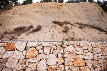 Close-up of a gabion wall made of irregular stones held by a metal mesh, set against a rugged...