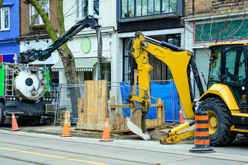 vacuum pump truck and backhoe at work site on city street with shop fronts in background shot in...