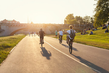 Obraz premium Scenic view youth people ride bicycles sunny Dresden Elbe riverside path against warm sunset glow active leisure. Rider perform wheelie beside historic Augustus bridge crowd relaxes grass lawn meadow