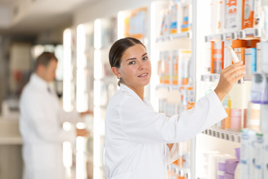 Positive young female pharmacist placing medical product on shelf in chemist's shop