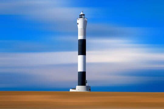   Tall black and white lighthouse on abstract beach with a bright blue and white sky, the beach and sky have motion in them whilst the light house is in sharp focus, Dungeness, Kent United Kingdom