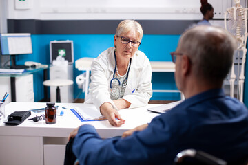 Obraz premium Caucasian old man listens as female physician explains his healthcare treatment. Retired male wheelchair user updating his elderly female doctor about his recovery progress in medical office.