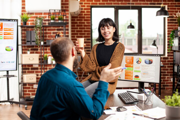 Smiling businesswoman with coffee, sitting on office desk and discussing with male coworker in...