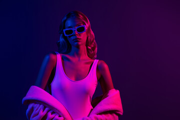 Chic young woman with blond hair in a bodysuit and sunglasses under neon pink light on dark purple...
