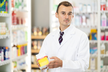 Fototapeta premium Smiling young man chemist demonstrating medicament in box in pharmacy store