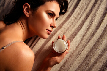 Elegant young woman holding a skincare product while lying on a textured grey surface, showcasing...