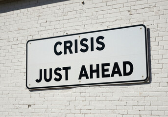 Crisis Just Ahead sign ominously warns of impending trouble against a stark brick wall background.