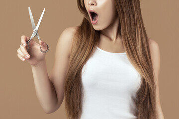 Obraz premium Shocked millennial woman with long straight hair holding scissors on brown studio background. Portrait of emotional young lady not willing to do haircut, opening mouth in surprise