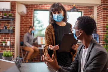 Caucasian female manager holding clipboard and advising african american male employee in startup office. Colleagues wearing face masks share ideas on business strategy in modern workspace.