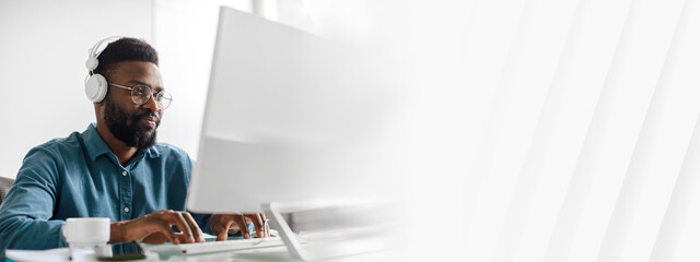 A young African American man works diligently at his desk in a modern office. He wears headphones...