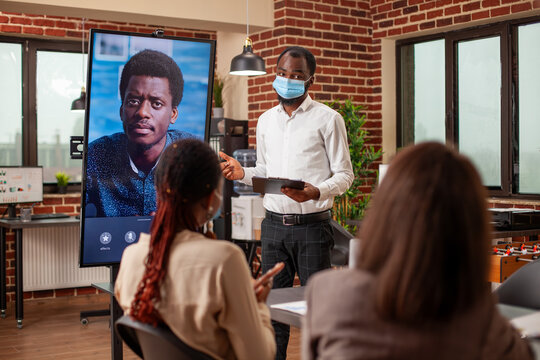 Black man in face mask gestures to screen with active videocall of african american client, providing project updates. Team engages in virtual meeting with remote manager, analyzing business metrics.