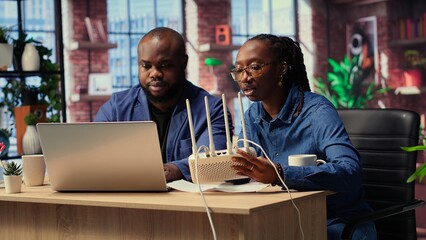 African American couple at home office troubleshooting their Wi Fi modem and discussing solutions to fix a weak connection affecting their remote work productivity. Router guide files. Camera B.