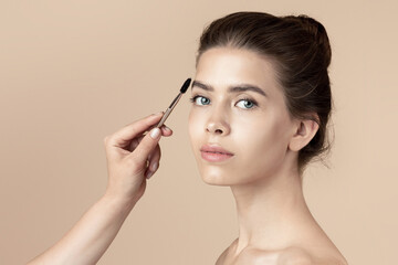 Makeup artist applying eyebrow mascara on young girl's face over beige background