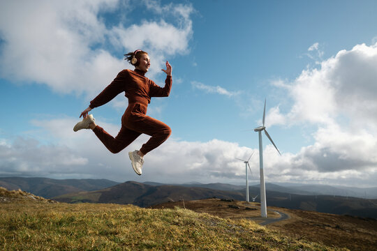 Young sportswoman jumping listening music with wind turbines and mountains in background
