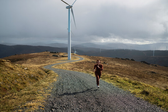 Young athlete running on a gravel road near wind turbines, enjoying nature and music