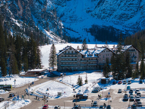 Aerial view of a snow covered alpine building near Braies Lake in the Dolomites, Italy, with evergreen trees, rocky peaks, and a parking area visible.