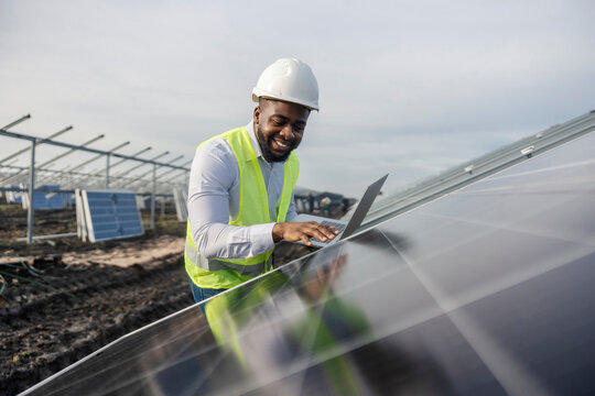 Smiling interracial engineer with laptop inspecting solar panels at solar farm.