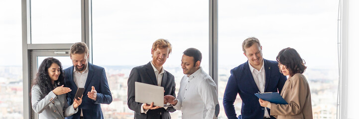 A group of business professionals are having a meeting in a modern office. They are standing around a large wooden table, looking at laptops and tablets, and discussing work.