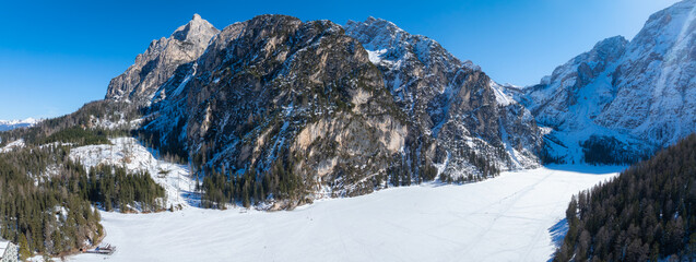 Aerial view of Braies Lake in winter, frozen and snow covered, surrounded by cliffs, rugged peaks,...