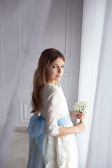 beautiful girl in elegant white dress with bouquet of flowers, looking out the window during first communion