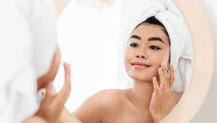 Cheerful asian young woman applying beauty product on her face, looking at mirror. Pretty smiling lady wrapped in towel using face cream after shower, sitting in front of mirror, face care concept