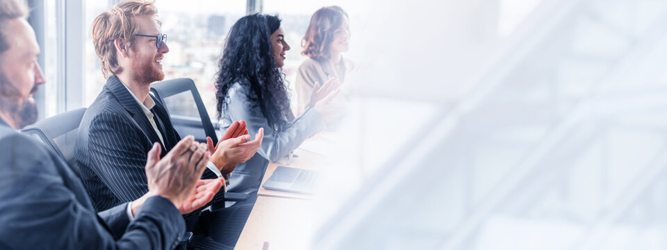 A group of business professionals sit at a table in a modern office, clapping their hands in appreciation as they listen to a presentation. They are dressed in formal attire