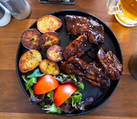Black ceramic plate contains furnace baked pork ribs, fried potatoes and pieces of fresh vegetables. Meat is complemented with tomato slices and arugula leaves..