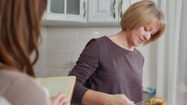 Close-up rear view of girl holding book while observing aunt cooking in bright kitchen, aunt lifts whisk with thick brown creamy pap, focused on preparation