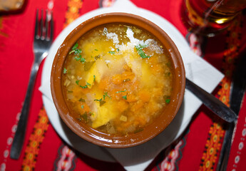 Deep clay bowl with Bulgarian fish and vegetable soup, complemented by cutlery and strong drink. Table is covered with red tablecloth with traditional Eastern European pattern.