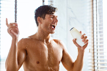 Attractive young wet man singing in the shower, holding shampoo bottle and using it as a microphone