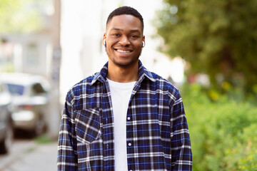 Fototapeta premium Portrait of happy black hipster wearing earbuds and posing at camera, spending time outdoor, selective focus