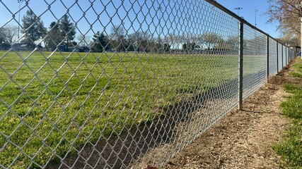 empty sports park enclosed by chain link fence under cloudy sky metal posts stretch along gravel path silent field beyond holds echoes of games past stillness rests on worn bleachers and grass
