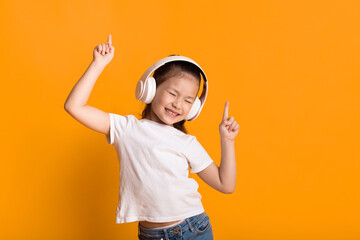 Good Mood. Adorable asian kid listening to music wearing wireless headset and dancing, isolated over yellow wall