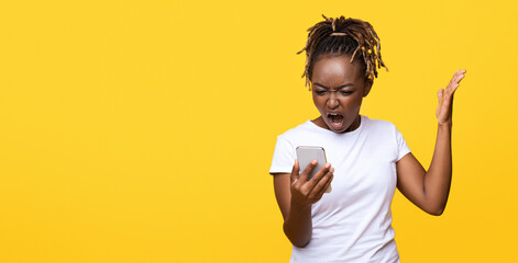 Furious black lady standing on yellow studio background, screaming at smartphone in her hand, gesturing, expressing anger, panorama with empty space. Unhappy african american lady holding mobile phone