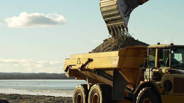 Excavator loading industrial dump truck at beach building site, slow motion