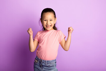 Yes. Happy little asian girl shaking fists, making winner gesture, standing at studio isolated over pastel purple wall