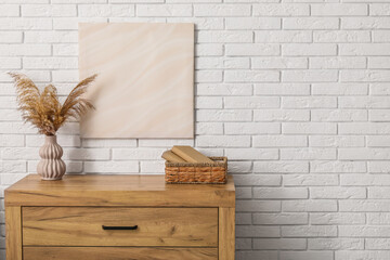 Chest of drawers with pampas grass, books in basket and picture on white brick wall in room. Closeup