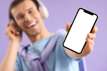 Young man in headphones with blank mobile phone on lilac background, closeup