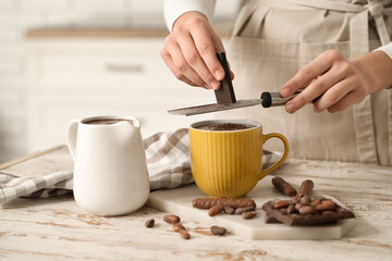 Woman grating bar of chocolate onto cup of melted chocolate on table in kitchen. Closeup