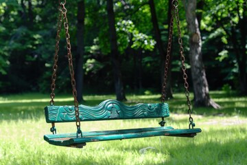 A rustic green swing hangs from chains in a lush, sunny forest, inviting relaxation amid the tranquil natural surroundings.