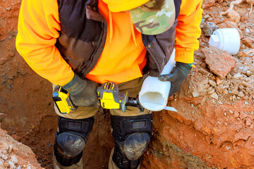 Construction worker in orange hoodie, gloves uses power tool to cuts pvc wastewater pipes in trench