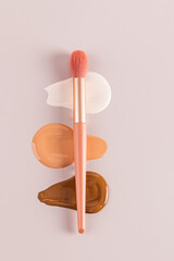 Three gorgeous strokes of liquid foundation in various tones on a beige vertical background with a professional makeup brush lying on them. top view
