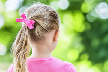 a child with blonde hair tied in two ponytails, wearing pink hair ties and a pink shirt. the background features a green, outdoor setting with a bokeh effect,