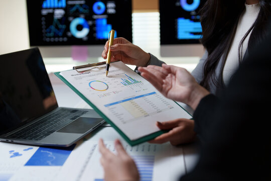 Two businesswomen analyzing a monthly budget report, pointing at charts and graphs on a clipboard, with a laptop and financial documents on the table in a modern office