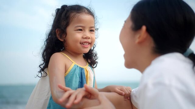 Happy asian mom applying sunscreen to her cute child&rsquo;s face on the beach for skin protection from the sun, a young mother carefully smears protective cream on her daughter&rsquo;s skin for safe outdoor fun.