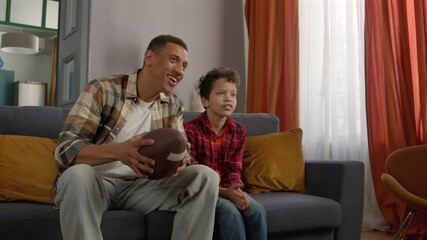Excited attractive African American father with American football ball and cheerful cute teenage son watching football game on tv, celebrating scoring drive and giving high five at home.