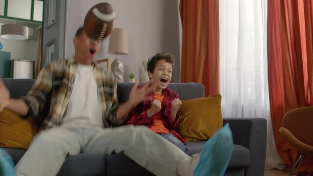 Excited school age African American boy and black father with football ball watching American football game on tv, embracing and emotionally celebrating touchdown in domestic room.