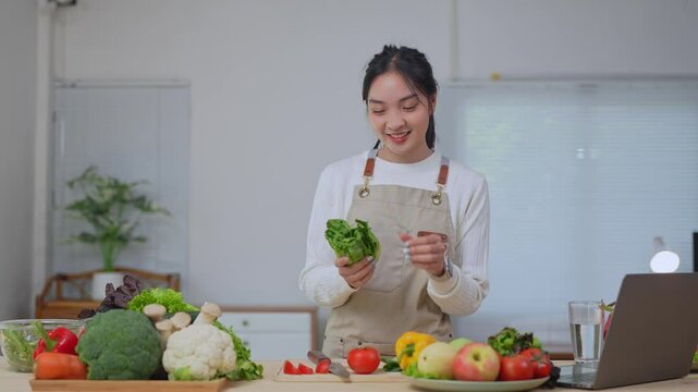 Young woman food blogger wearing an apron is showing lettuce while recording a video tutorial with her laptop in a kitchen full of fresh vegetables and fruits