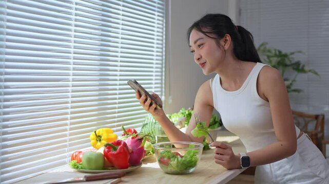 Young woman stands at her kitchen counter, illuminated by natural light filtering through window blinds, preparing and eating a salad while engaging in phone calls and browsing her smartphone