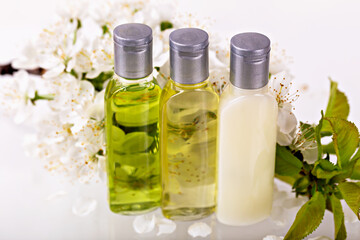 Three transparent bottles with silver caps containing skincare oils and lotion are placed among fresh white blossoms. Natural aesthetic emphasizes organic beauty, wellness and self-care concepts